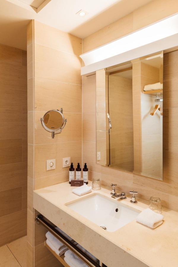 Townhouse Dresden Hotel Exterior photo The photo shows a modern bathroom with light beige tiles and a clean aesthetic. It features a sink area with a countertop, a large mirror mounted above the sink, and a round magnifying mirror attached to the wall. On the countertop, there are several